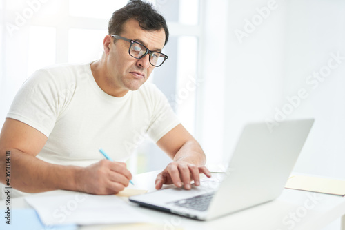 Work from home. Latin man in white t shirt and eyeglasses looking focused while sitting at the table, using laptop and making notes. Bright light coming from the window