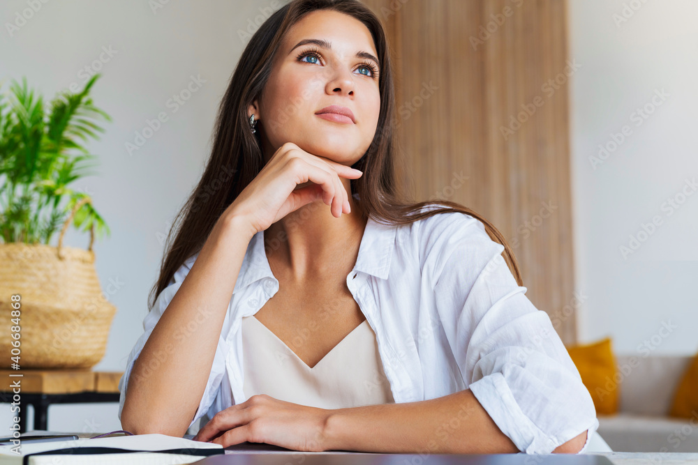 Beautiful young business woman is sitting at table of closed laptop. Thoughtful girl, smiles, thinks about pleasant things. Girl in white shirt is imagining and planning events. Work from home Beautiful young business woman is sitting at table of closed laptop. Thoughtful girl, smiles, thinks about pleasant things. Girl in white shirt is imagining and planning events. Work from home