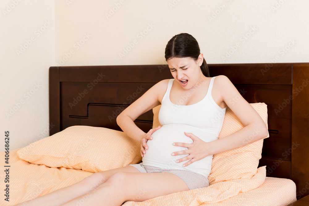 Young pretty pregnant woman, in pain, sitting on bed in bedroom, preparing to give a birth Young pretty pregnant woman, in pain, sitting on bed in bedroom, preparing to give a birth