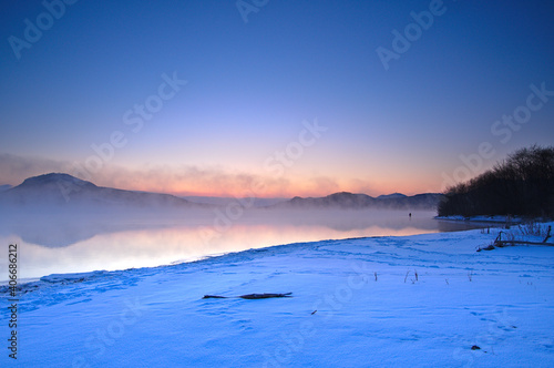 朝靄の漂う冬の朝の湖、雪に覆われた湖岸。夜明けの空のグラデーションと湖岸の森のシルエット。阿寒摩周国立公園の屈斜路湖。北海道、日本。