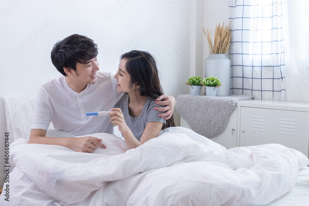 Happy young Asian couple looking and smiling together and wife holding pregnancy test in hand on bed in white bedroom. Happy young Asian couple looking and smiling together and wife holding pregnancy test in hand on bed in white bedroom.