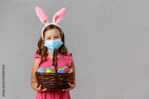 Happy cute little girl in the ears of the Easter bunny in a protective medical mask on a gray background. The child holds a basket with colorful eggs.