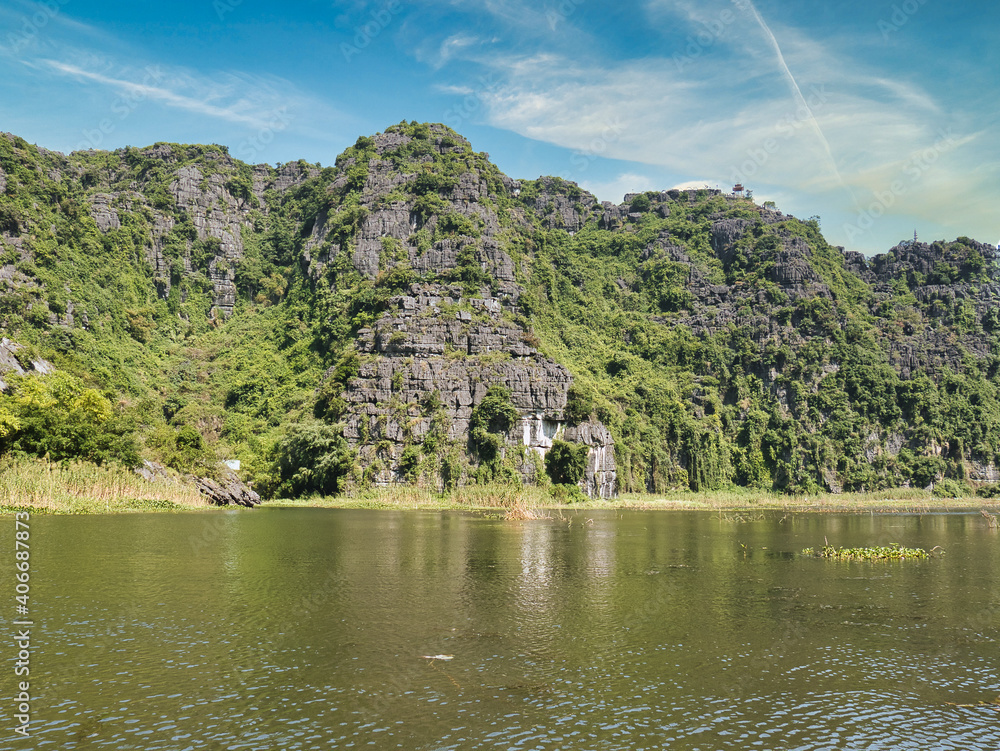 Kalksteinberge mit Wald an einem sonnigen Tag, vom Fluss aus gesehen, in Trang An, Tam Coc, Ninh Binh, Vietnam. Das Gebiet trägt den Spitznamen "die Ha Long Bay im Landesinneren". - obrazy, fototapety, plakaty 