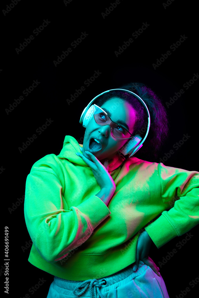Singer. African-american woman isolated on dark studio background in multicolored neon. Listening to music with headphones. Concept of human emotions, facial expression, sales, ad, fashion. Singer. African-american woman isolated on dark studio background in multicolored neon. Listening to music with headphones. Concept of human emotions, facial expression, sales, ad, fashion.