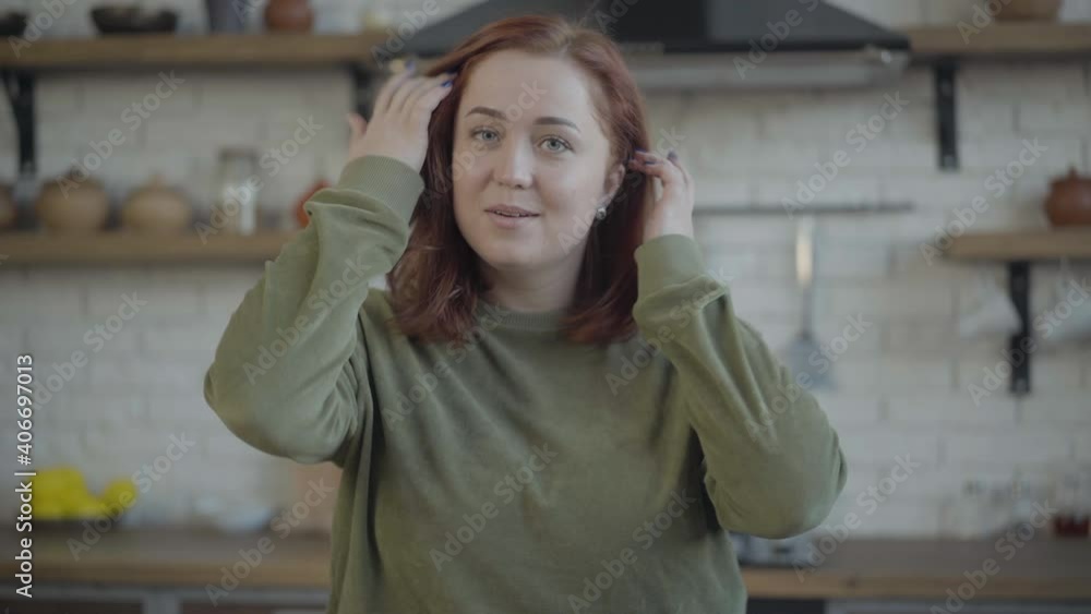 Middle shot of redhead young Caucasian woman with excited facial expression looking at camera. Portrait of cheerful happy chubby lady posing in kitchen at home indoors. Middle shot of redhead young Caucasian woman with excited facial expression looking at camera. Portrait of cheerful happy chubby lady posing in kitchen at home indoors.