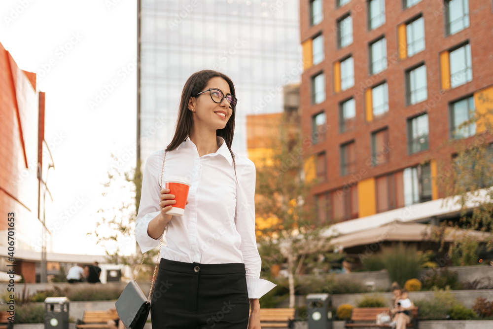 Businesswoman having a break from work and relaxing while walking with a coffee to go with business center in background.