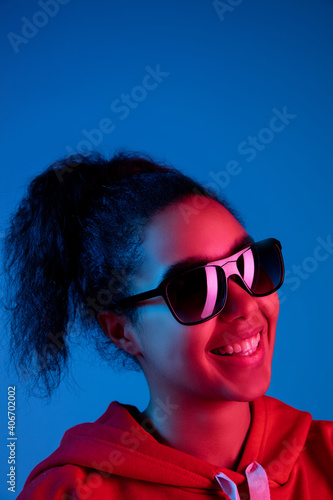 Laughting. African-american woman's portrait on blue studio background in red-pink neon light. Sunglasses wearing. Concept of human emotions, facial expression, sales, ad, fashion. Copyspace.