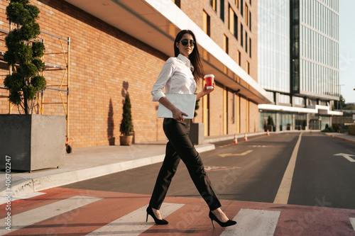 Businesswoman having a break from work and relaxing while crossing the street on zebra holding coffee to go with business center in background.