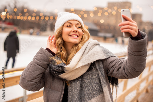 Woman doing selfie on the background of festive Christmas ice rank. Lady wearing winter warm knitted clothes.