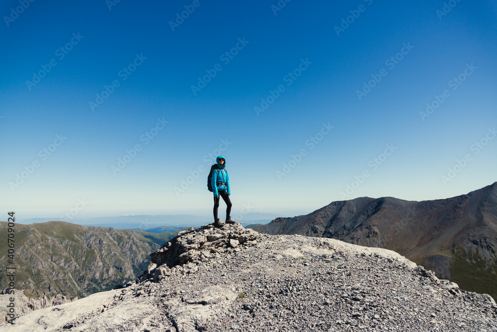 Woman traveler with a backpack in a hood stands on the top of a mountain against the backdrop of a cloudless blue sky. Social distancing concept when traveling