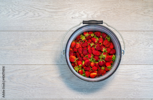Overhead shot of a tin pail filled to the top with fresh strawberries, on a wooden planked table, painted in soft white. Copy space to right of frame.