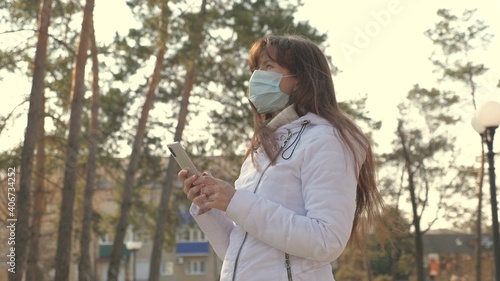 A young woman in medical protective mask stands with smartphone on city street in park. Coronavirus protection. Tourist on street put on protective masks against viruses. health and safety. covid-19