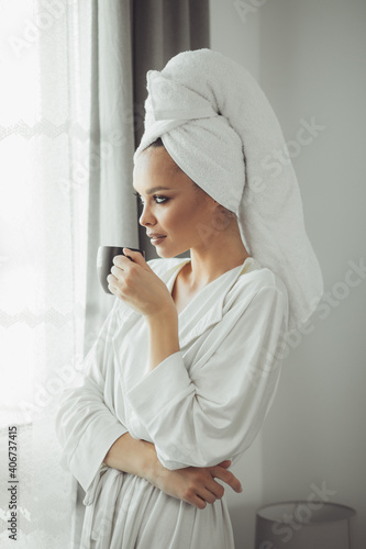 Young woman in bathrobe drinking coffee and looking through the window.