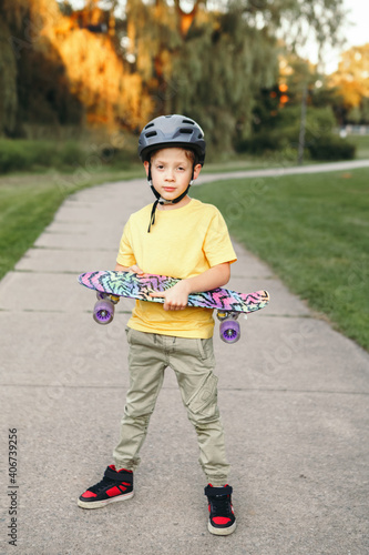 Happy Caucasian boy in grey helmet standing with skateboard on road in park on summer day. Seasonal outdoor children activity outdoors sport. Healthy childhood lifestyle.