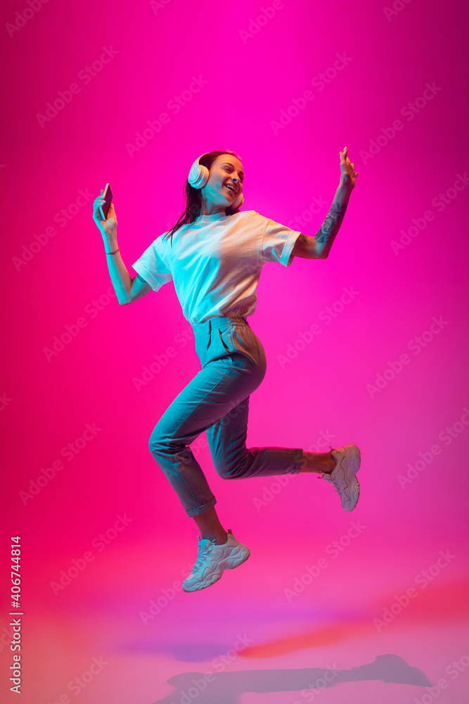 Jumping high. Caucasian woman's portrait isolated on pink studio background in mixed neon light. Listening to music with phone. Concept of human emotions, facial expression, sales, ad, fashion