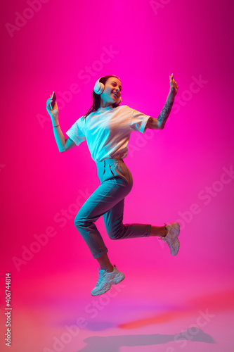 Jumping high. Caucasian woman's portrait isolated on pink studio background in mixed neon light. Listening to music with phone. Concept of human emotions, facial expression, sales, ad, fashion