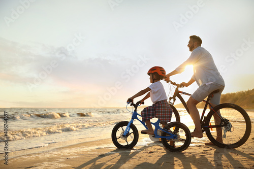 Happy father with son riding bicycles on sandy beach near sea at sunset