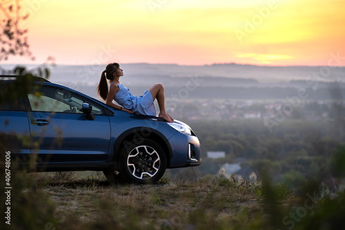 Happy young woman in blue dress sitting on her vehicle bonnet looking at sunset view of summer nature. Travelling and vacation concept.