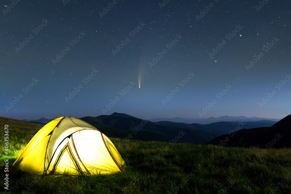 Tourist hikers tent in mountains at night with stars and Neowise comet with light tail in dark night sky.
