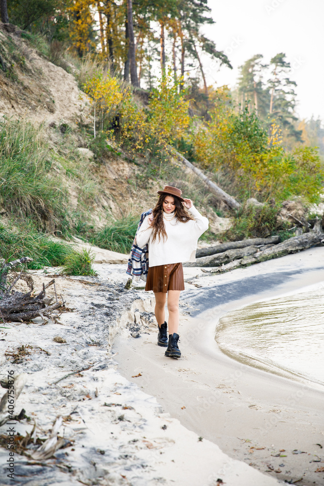young beautiful girl in a white sweater, brown skirt and brown hat are walking on the coast  near the lake