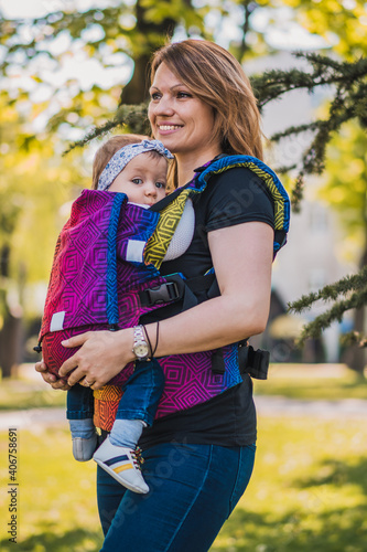 Fototapeta Naklejka Na Ścianę i Meble -  Beautiful happy young mother with baby carrier in park. Mom walking with infant in baby carrier. Happy mother carrying her child by ergonomic baby carrier.