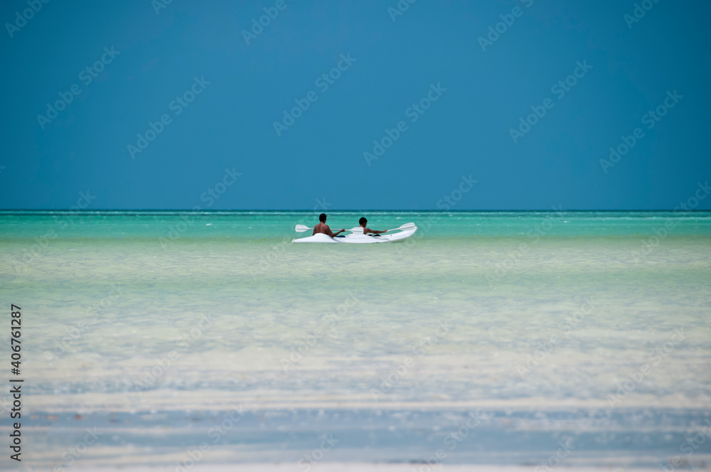 Two unrecognizable men sailing a kayak in the Caribbean Sea near a tropical beach on the island of Holbox, Mexico. In the background the blue sky