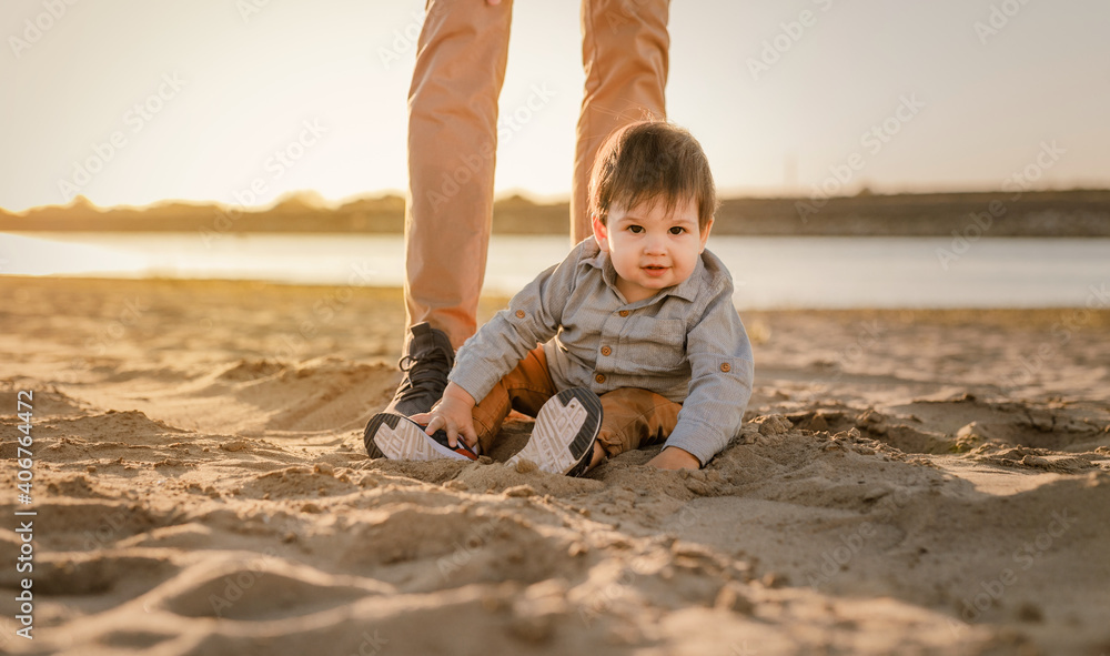 One-year-old boy sits on his father's feet in the sand on the riverbank One-year-old boy sits on his father's feet in the sand on the riverbank