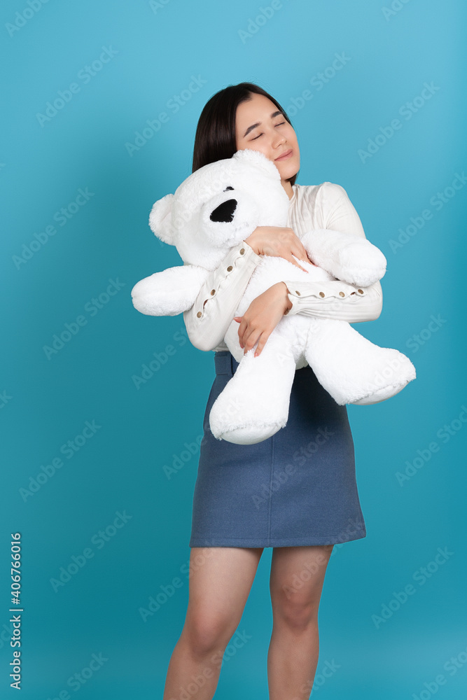 charming young Asian woman with her eyes closed tightly hugs a large white teddy bear valentine's day gift isolated on a blue background. charming young Asian woman with her eyes closed tightly hugs a large white teddy bear valentine's day gift isolated on a blue background.