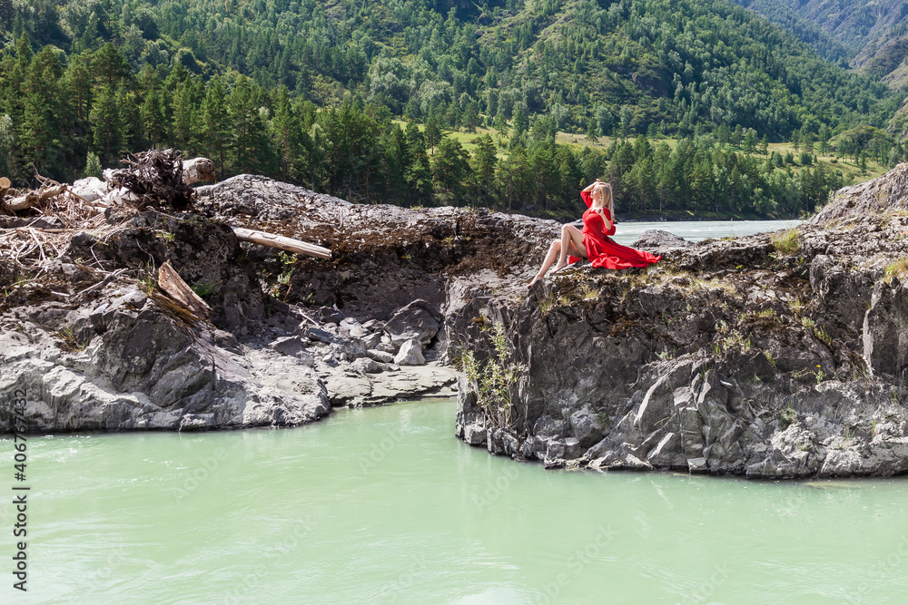 Blonde woman in red vintage dress holding up arms, sits stretching her long legs on the stones on banks of  Katun river against background of mountains.