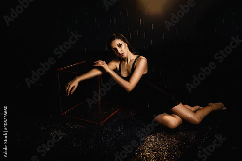 A slender brunette in a black dress on a black background in the studio under drops of rain, illuminated by spotlights
