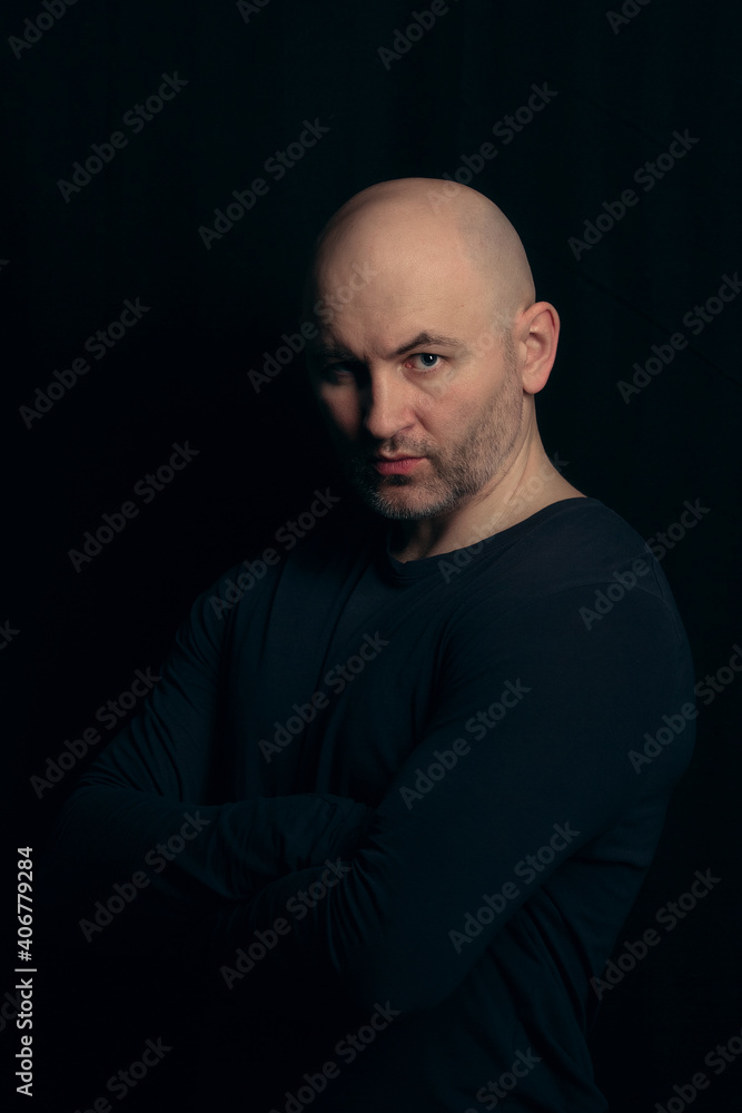 Portrait of a brutal bald man in a dark style on a black background. A bald athletic guy in a black turtleneck with a confident and bold look.