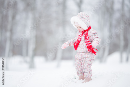 One baby girl in pink overalls and red scarf standing on white snow and enjoying beautiful winter day after blizzard at park. Side view.