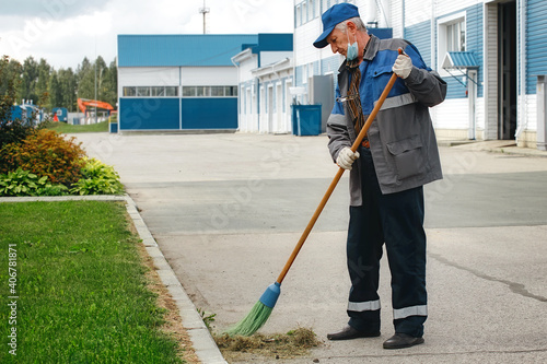 This is an elderly janitor with a broom in a medical mask on the street sweeping the territory. An old man in a work uniform works during a time of pandemic and unemployment.