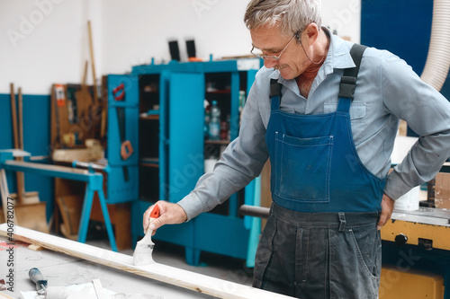 Bild auf Leinwand An elderly cabinetmaker in overalls and glasses paints a wooden board with a brush on a workbench in a carpentry shop