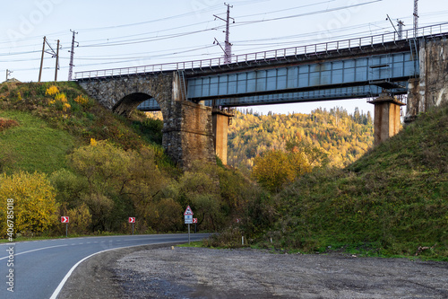 An old stone arched railway bridge over a road in the mountainous part of the Ukrainian Carpathians. Autumn mountain landscape - yellowed and reddened autumn trees combined with green needles.