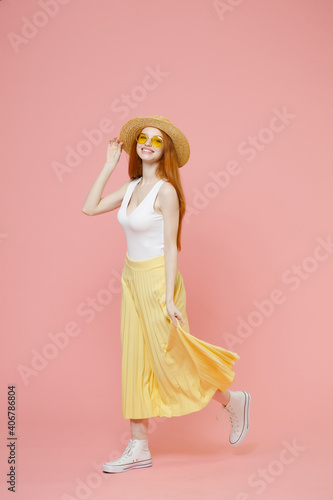 Full length body young smiling redhead ginger woman in straw hat glasses summer clothes maxi skirt walk going waving hand greet someone look camera isolated on pastel pink background studio portrait