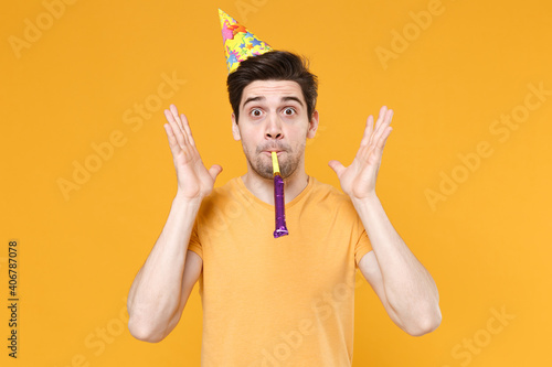 Canvas Print Young unshaved cute happy fun friendly caucasian man 20s in casual t-shirt wearing birthday hat cone blow pipe celebrating greeting spreading hands isolated on yellow color background studio portrait