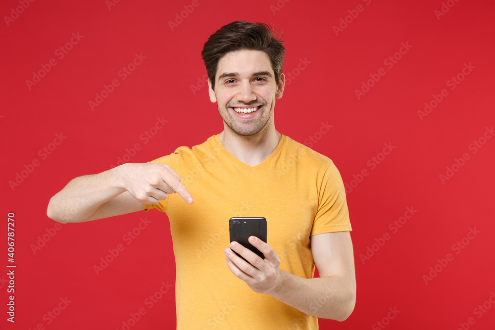 Young smiling unshaved caucasian man 20s in casual basic yellow t-shirt holding pointing index finger on modern mobile cell phone reading news isolated on red color background studio portrait. Young smiling unshaved caucasian man 20s in casual basic yellow t-shirt holding pointing index finger on modern mobile cell phone reading news isolated on red color background studio portrait.
