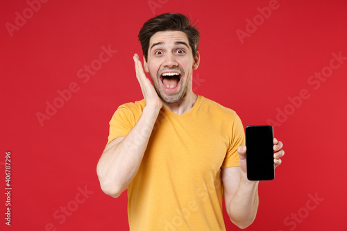 Young shocked unshaved man in casual basic design yellow t-shirt hold mobile cell phone with blank screen workspace copy space mock up spreading hand isolated on red color background studio portrait.