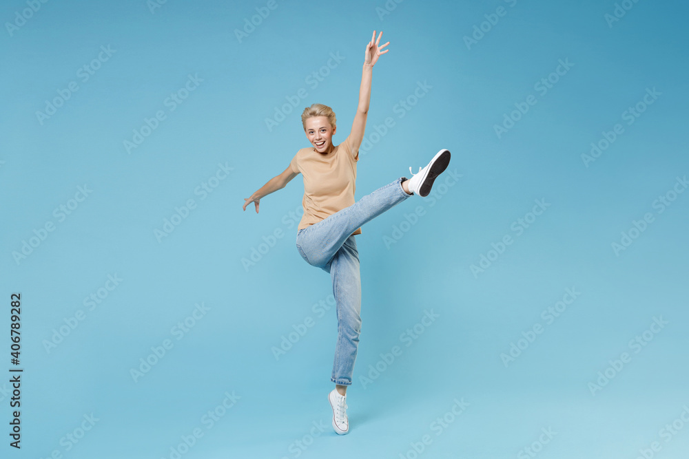 Full length of smiling fun happy caucasian young blonde woman 20s short haircut wear casual beige t-shirt jeans standing with raised up leg fooling around isolated on blue background studio portrait