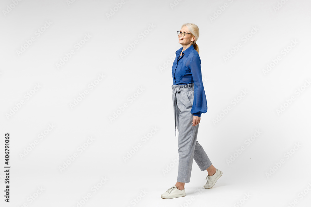 Full length side view blonde pensive happy successful employee business woman 40s in blue classic shirt gray pants glasses formal clothes walking going isolated on white background studio portrait. Full length side view blonde pensive happy successful employee business woman 40s in blue classic shirt gray pants glasses formal clothes walking going isolated on white background studio portrait.