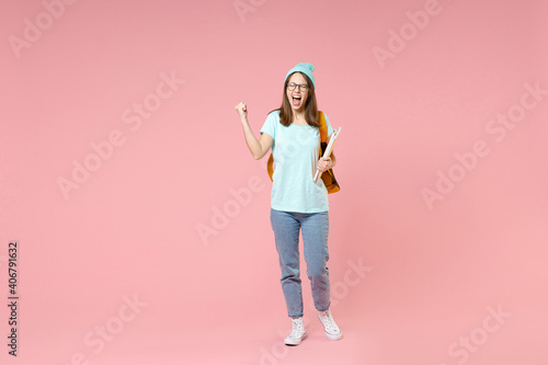 Full length portrait screaming young woman student in t-shirt hat glasses backpack hold notebooks doing winner gesture isolated on pink background. Education in high school university college concept.