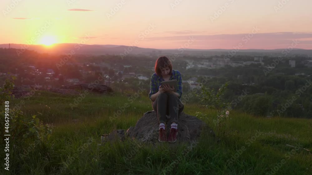 Brunette woman with a tablet pc sits on a stone among the grass on a mountain at sunset. Freelancer checks previously done work with concentration. Brunette woman with a tablet pc sits on a stone among the grass on a mountain at sunset. Freelancer checks previously done work with concentration.