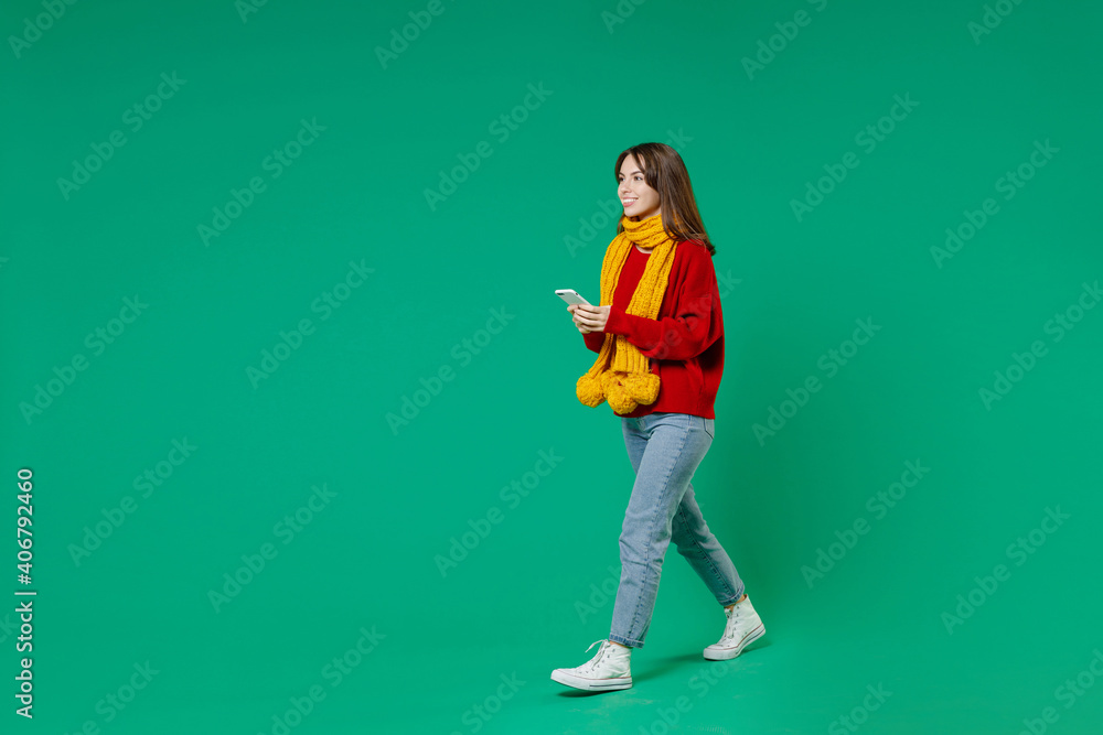 Full length side view of smiling cheerful young brunette woman 20s in knitted red sweater yellow scarf using mobile cell phone typing sms message isolated on green color background studio portrait. Full length side view of smiling cheerful young brunette woman 20s in knitted red sweater yellow scarf using mobile cell phone typing sms message isolated on green color background studio portrait.