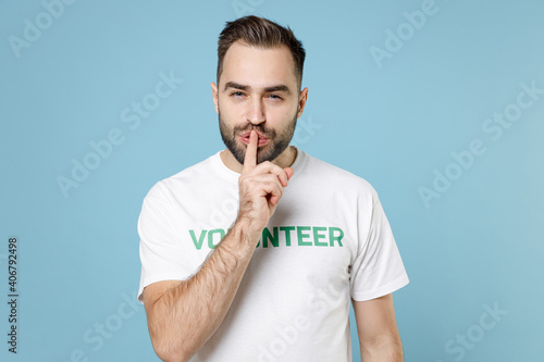 Secret young man in white volunteer t-shirt say hush be quiet with finger on lips shhh gesture isolated on blue background studio portrait. Voluntary free work assistance help charity grace concept.