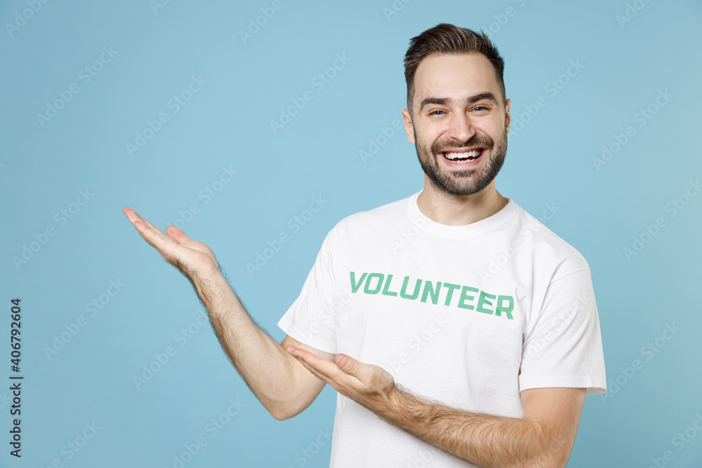Cheerful young bearded man in white volunteer t-shirt pointing hands aside on mock up copy space isolated on blue background studio portrait. Voluntary free work assistance help charity grace concept. Cheerful young bearded man in white volunteer t-shirt pointing hands aside on mock up copy space isolated on blue background studio portrait. Voluntary free work assistance help charity grace concept.