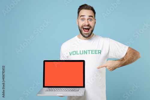Konstfotografi Excited young man in volunteer t-shirt point index finger on laptop pc computer with blank empty screen isolated on blue background studio