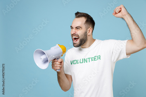 Fotografie Crazy young man wearing white volunteer t-shirt screaming in megaphone clenching fist isolated on blue color background studio portrait