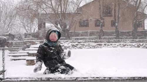 
little boy shakes off snow from steps of snow-covered staircase leading to garden and to thouse by river during snowfall photo