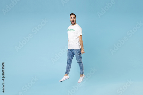 Full length of excited funny young bearded man 20s wearing white volunteer t-shirt jumping isolated on blue color background studio portrait. Voluntary free work assistance help charity grace concept.